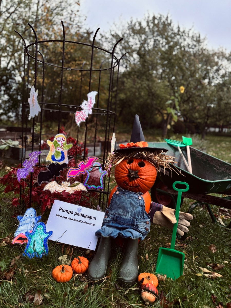 Pumpa med hatt, kropp i snickarbyxor och gummist&ouml;vlar sitter utomhus omgiven av pysslade sp&ouml;ken och fj&auml;rilar. En skylt med text: "Pumpapedagogen - Med r&auml;tt st&ouml;d kan all blomstra!"