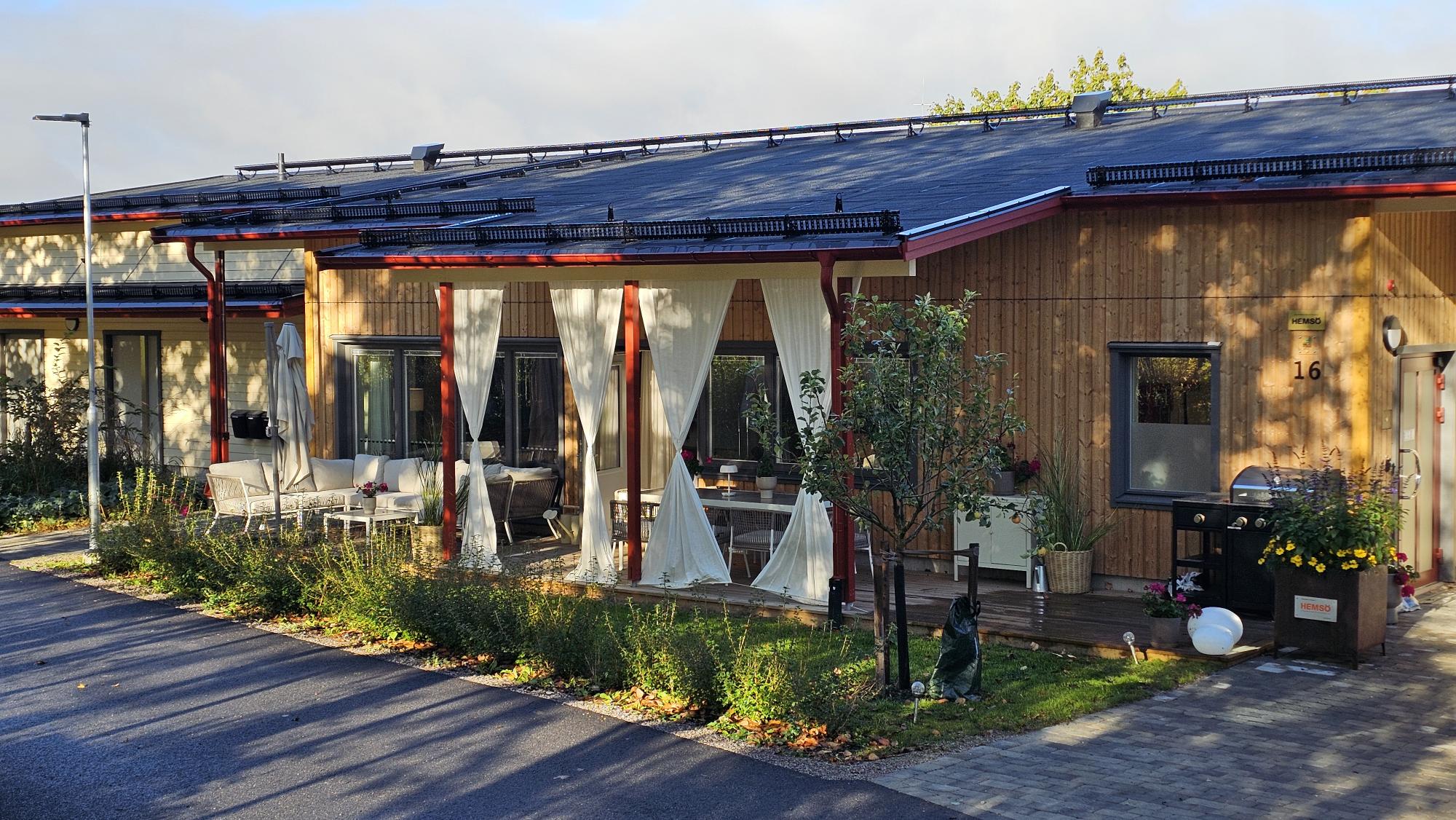 En modern villa med träterrass, solpaneler på taket och utemöbler. En liten trädgård med buskar och ett olivträd syns framför huset