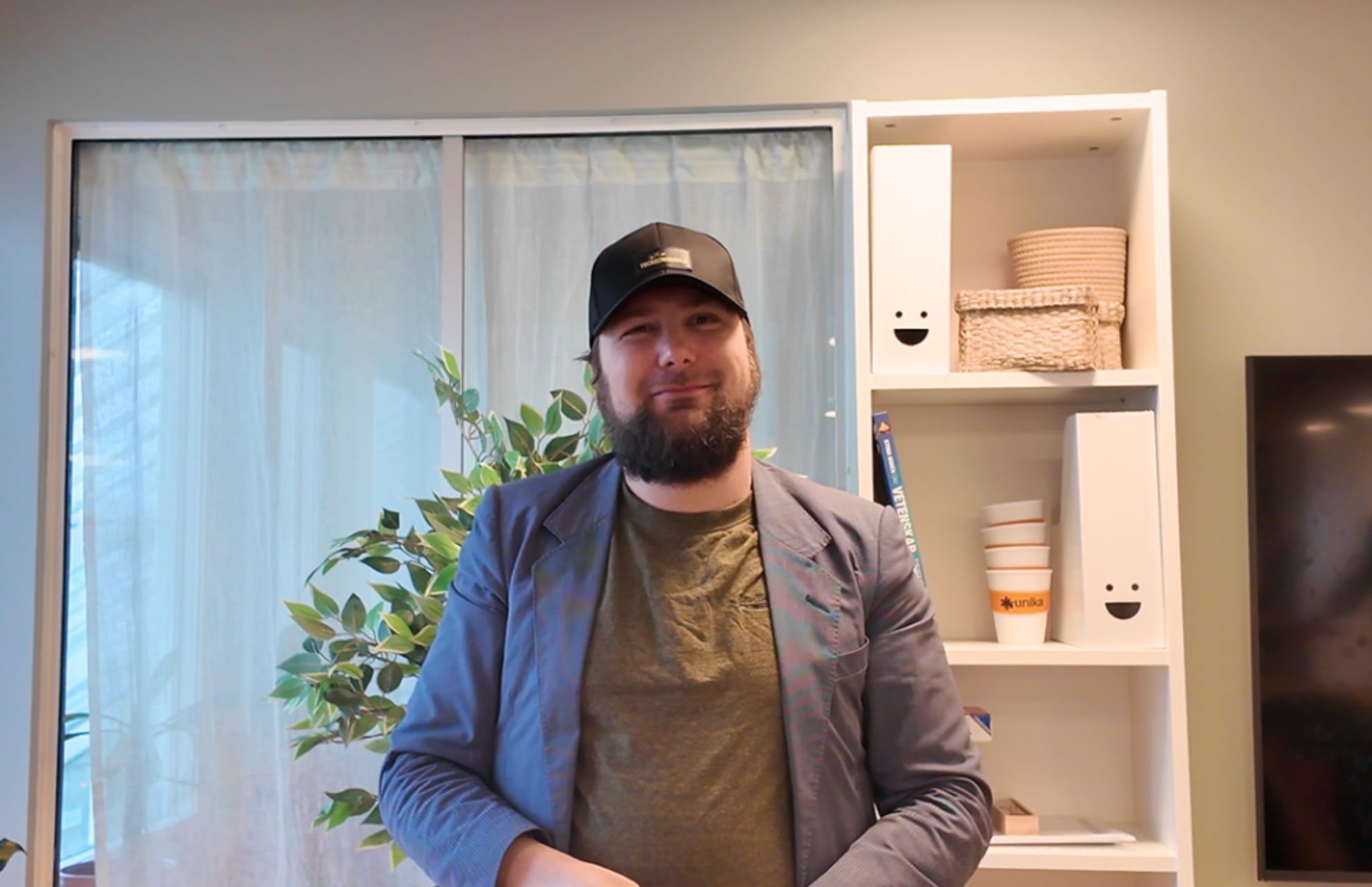 Oleg, med kavaj och keps, står i Unika Bergshamras vardagsrum